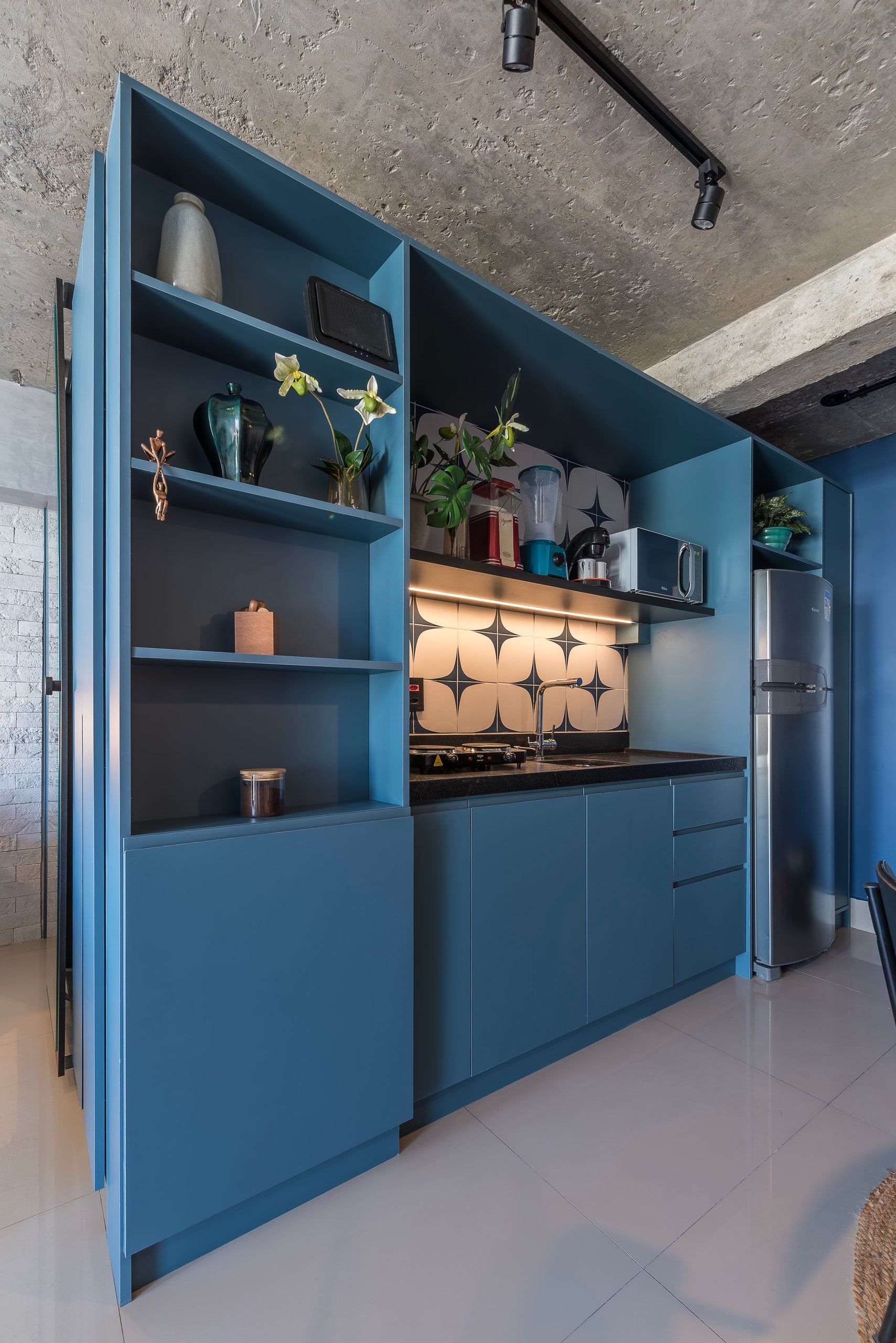 Cozinha compacta em marcenaria azul-petróleo, com prateleiras decoradas, revestimento geométrico preto e branco e teto de concreto aparente