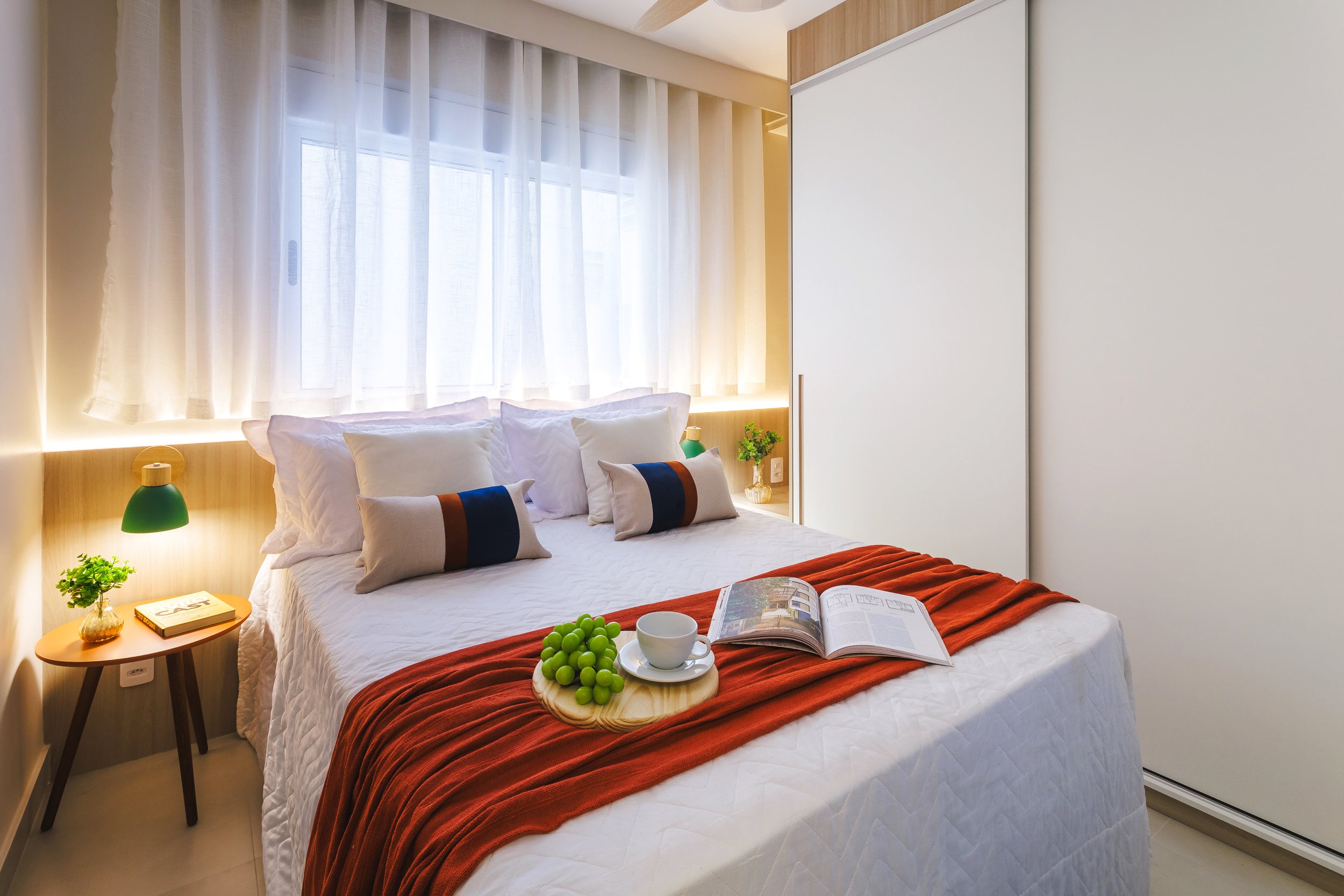 Cama de casal com roupa de cama branca e manta terracota, junto à cortina de linho