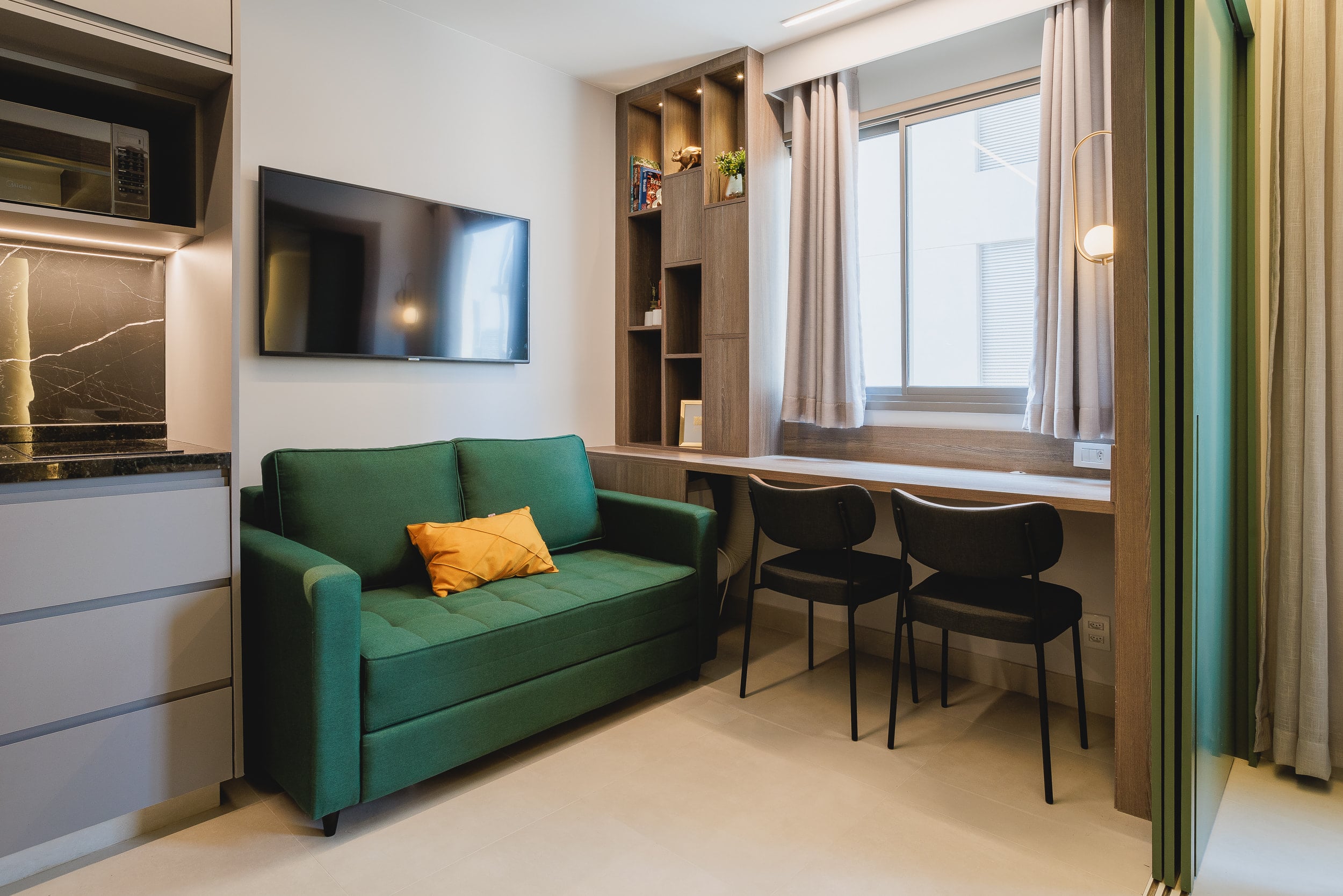 Studio em modo estar: sofá verde, bancada de trabalho e janela ampla