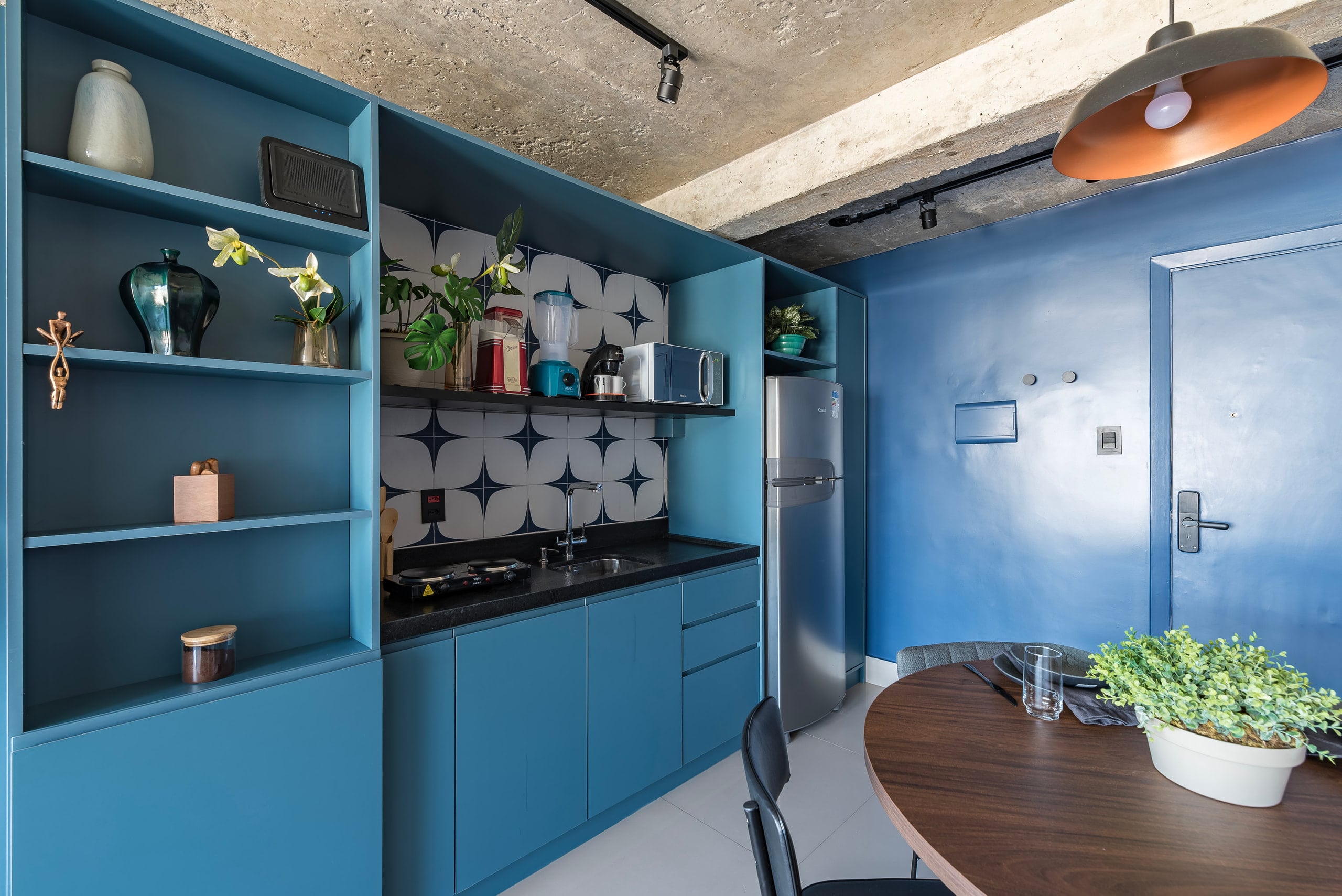 Cozinha em marcenaria azul com cooktop, prateleiras e teto de concreto