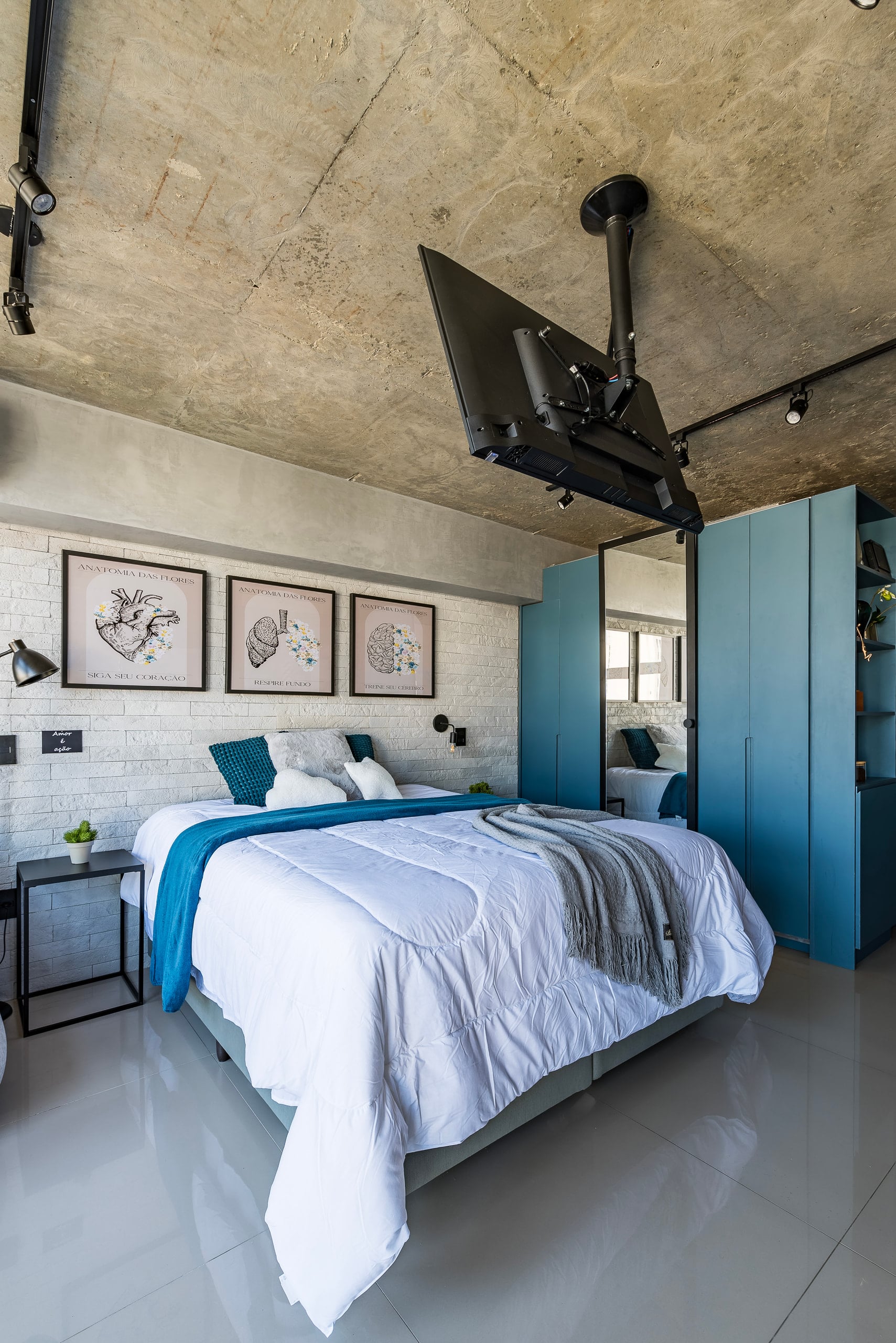 Cama com quadros na parede de concreto e trilho de spots no teto