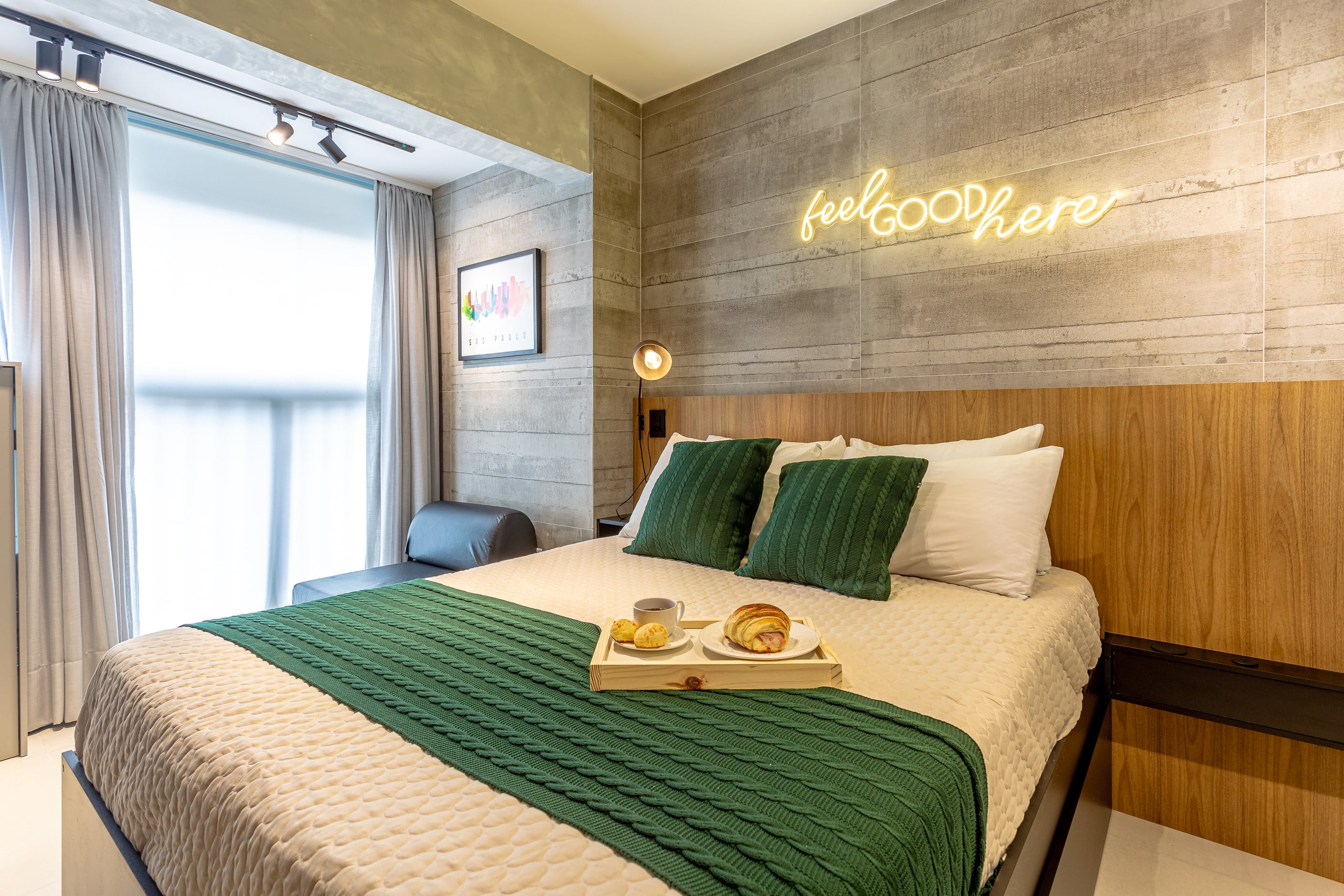 Cama junto à janela, com o letreiro “feel good here” na parede de concreto