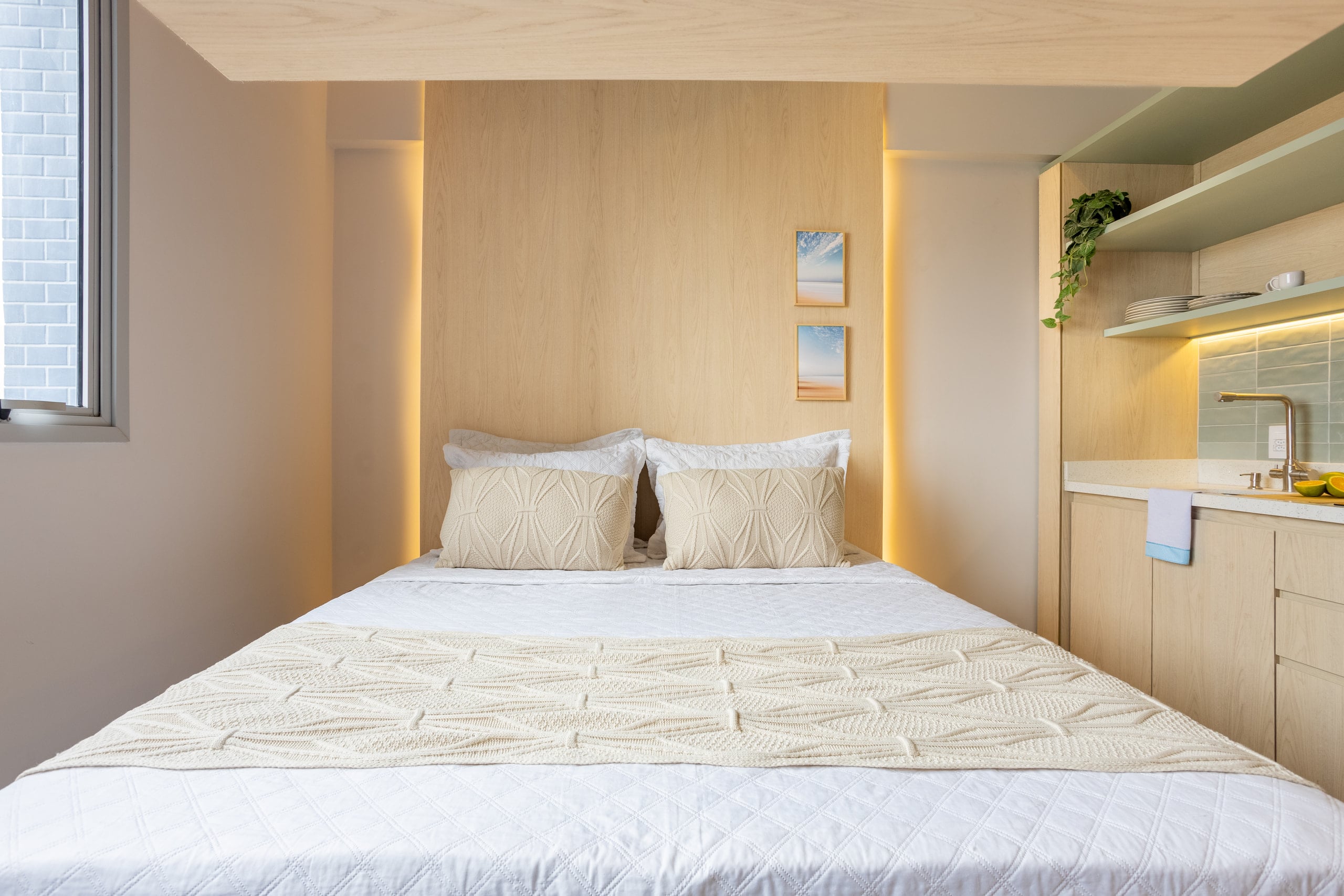 Cabeceira iluminada com quadros de paisagem e roupa de cama branca